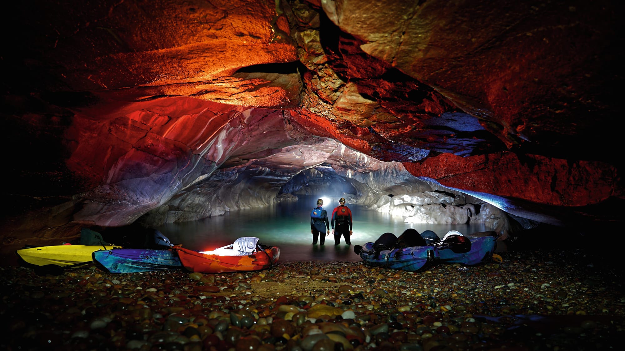 Kayaks are shored on a stony beach in an underground cave as two people explore the water with head torches. 