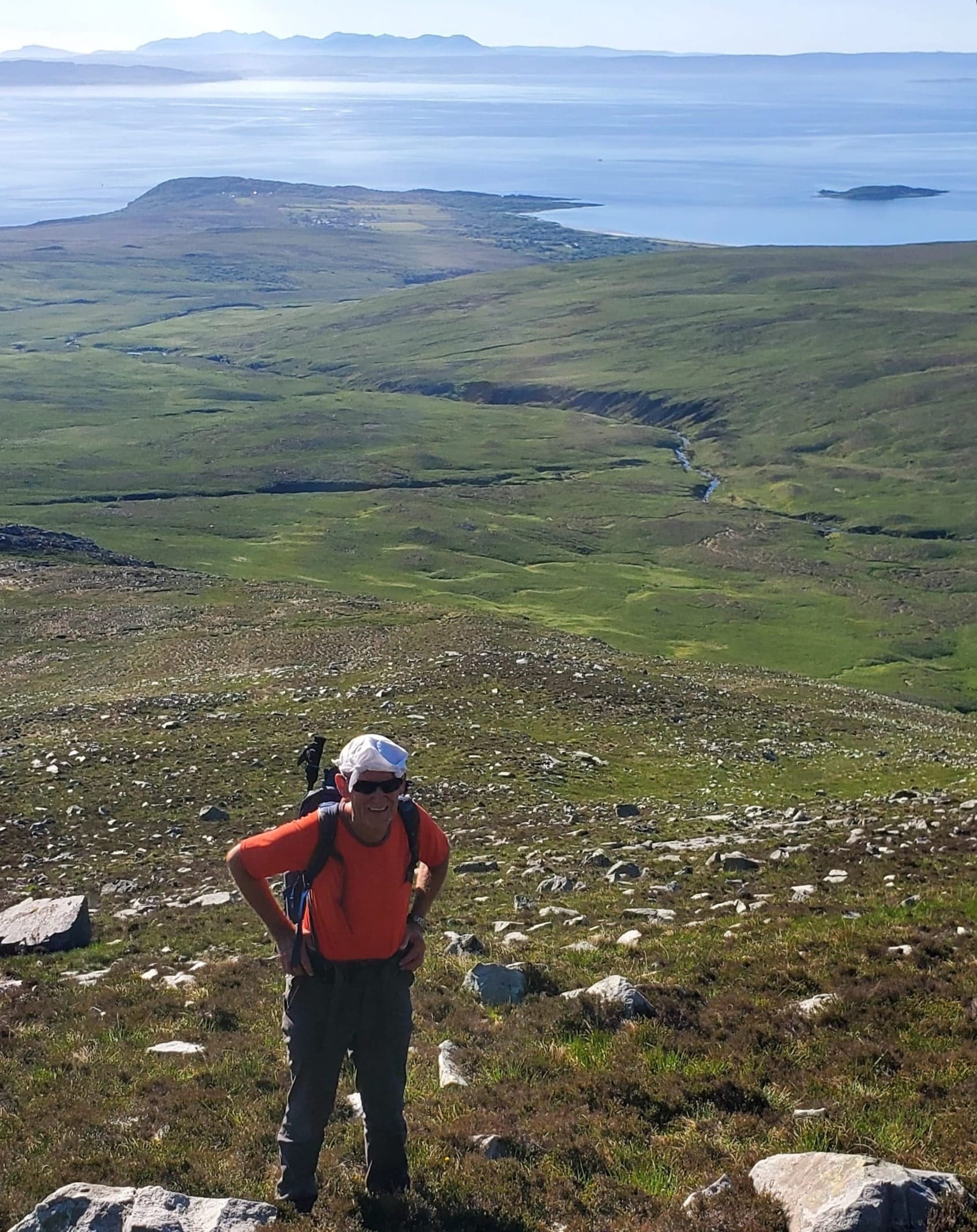 Ron wearing hiking gear standing on a green hill, the sea in the background.