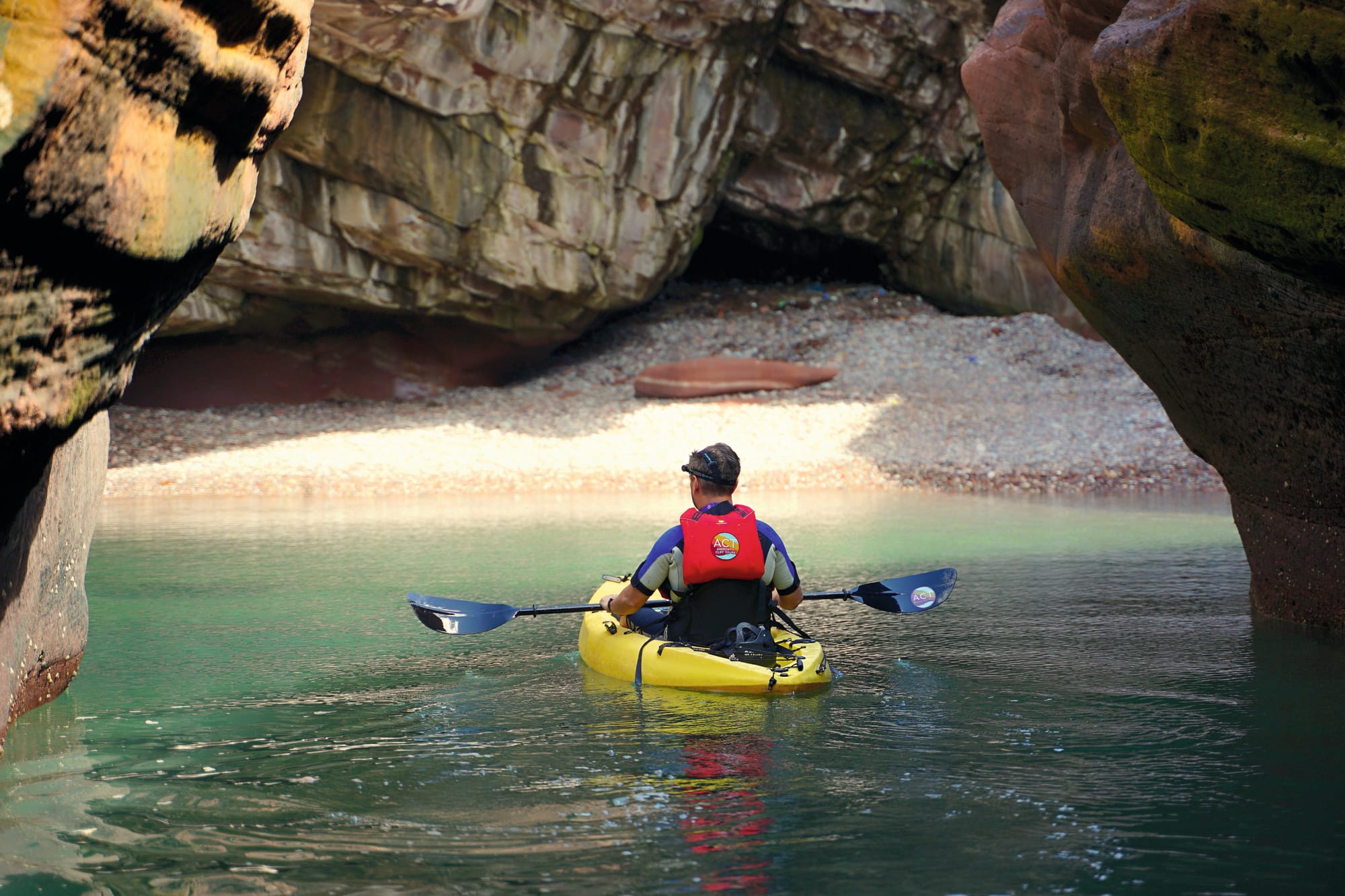 A man kayaking out of a cave towards a small beach. 