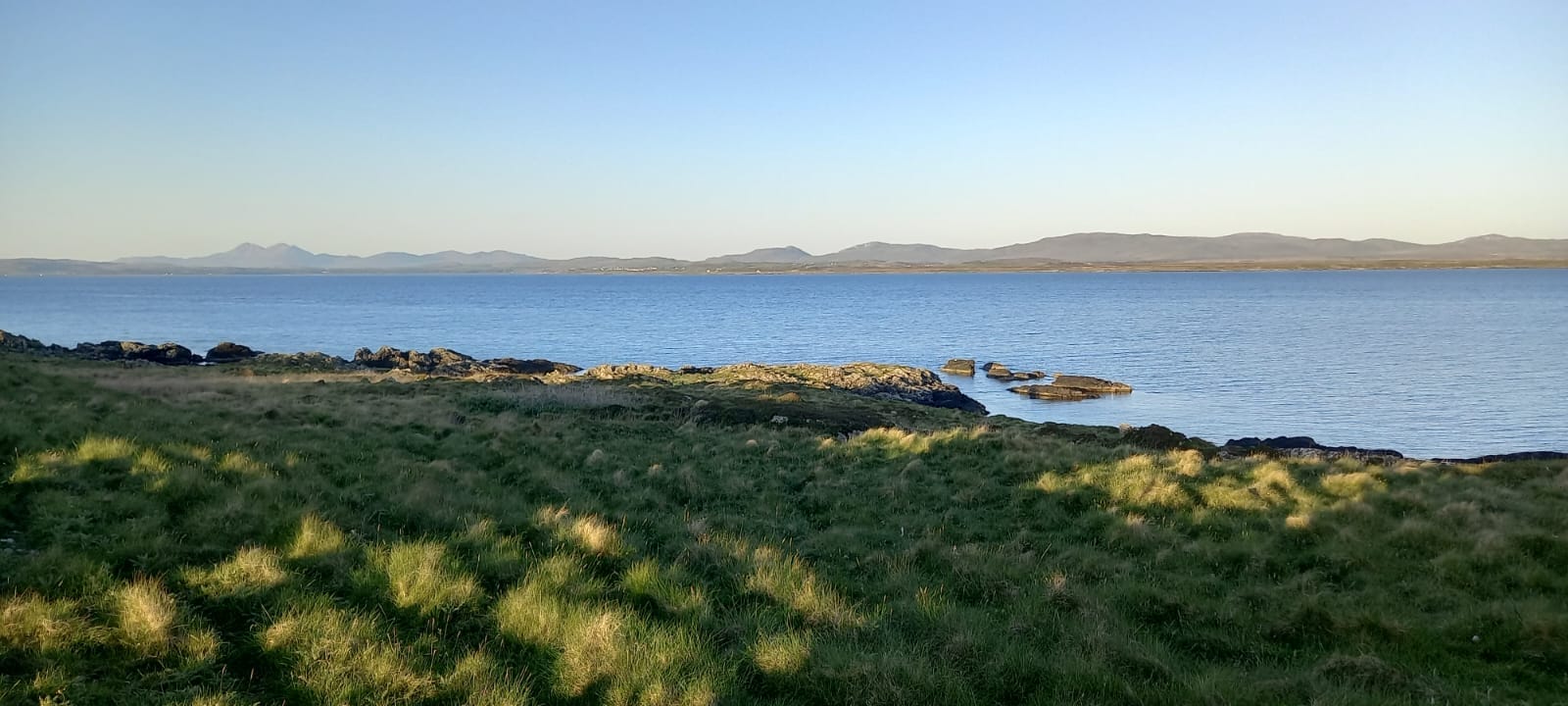 Sun-dappled grass in the foreground, looking across the water to distant hills.