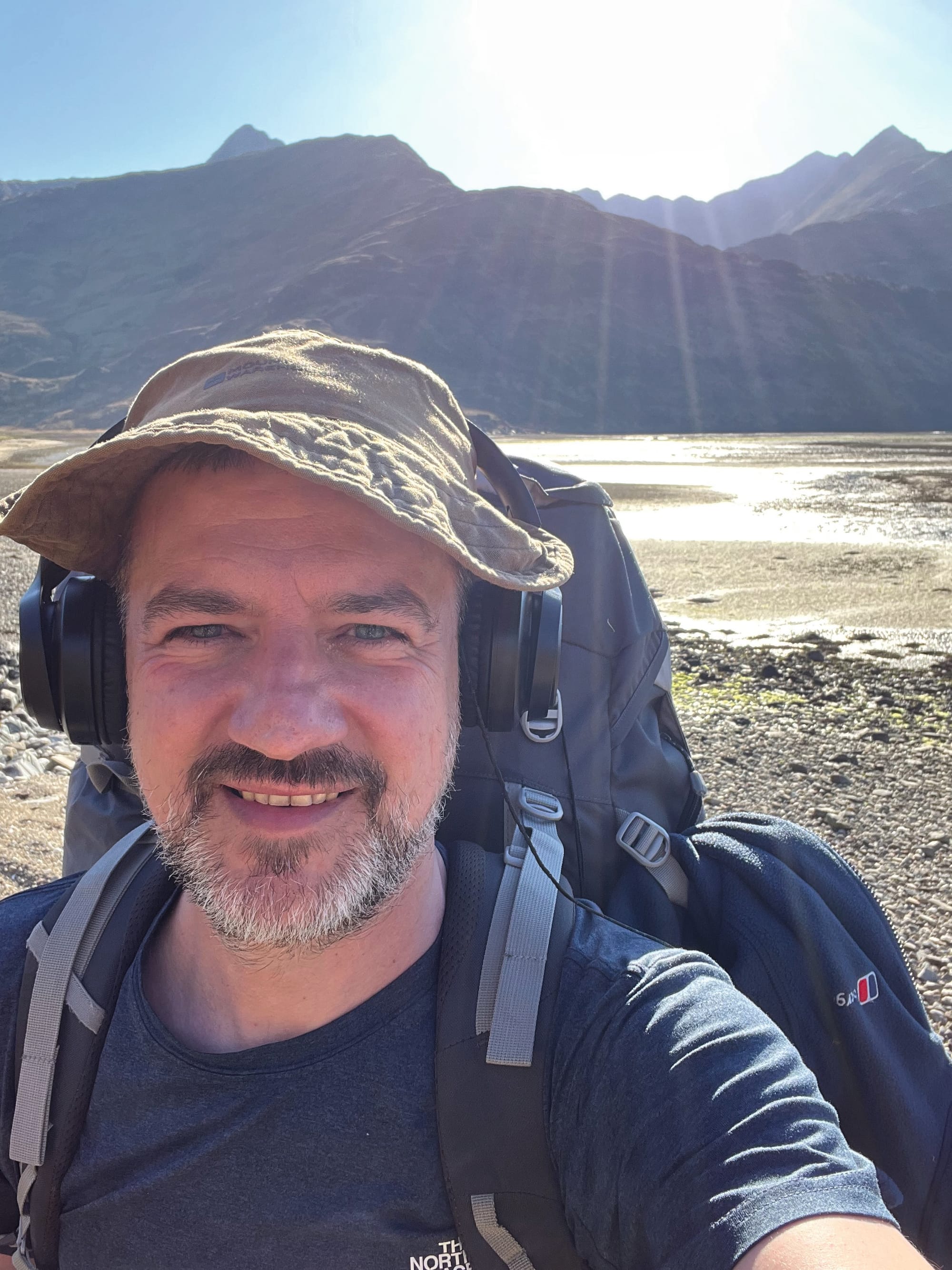 John smiles for a selfie wearing a hat, headphones and his hiking rucksack. The sun shines from behind him.