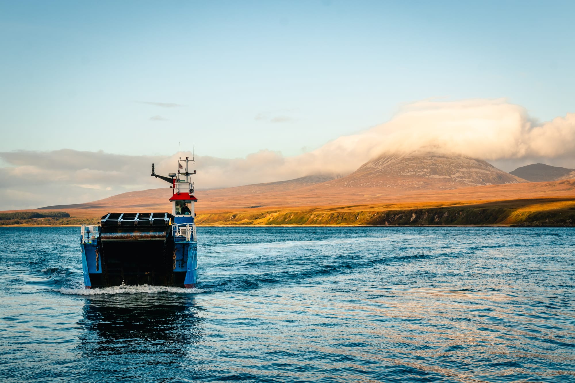 A small ferry sails to the golden hills of Jura which are topped with fluffy clouds.