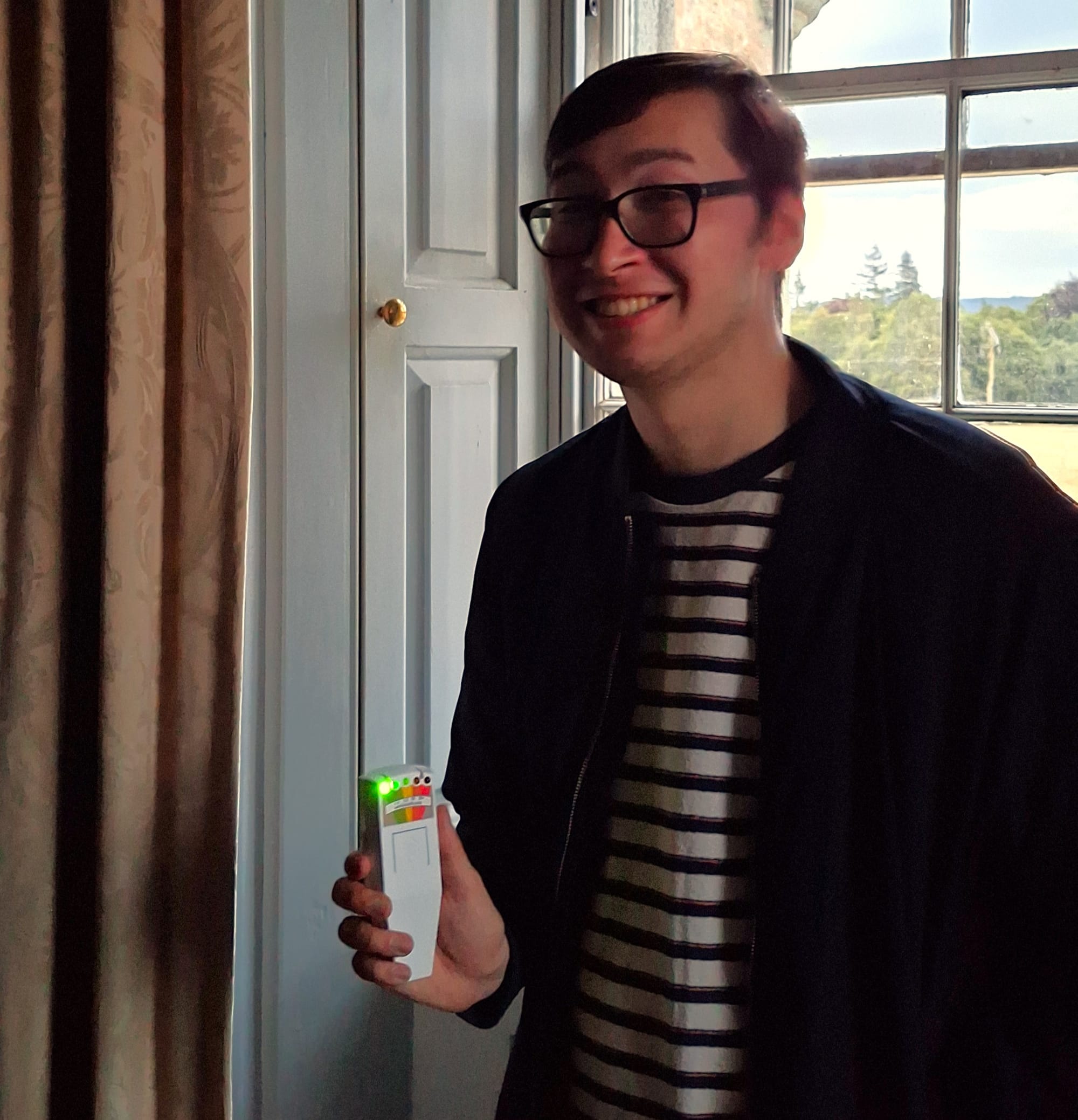 James stands by a window in the castle and holds up the EMF reader, smiling.