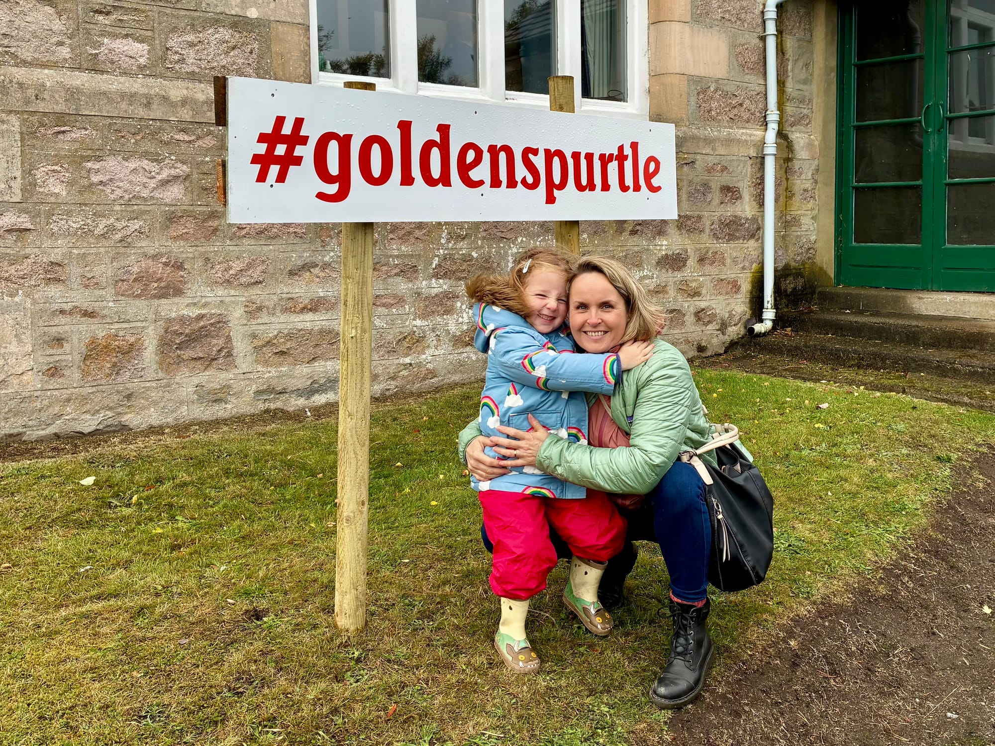 A woman with short blond hair crouches down and cuddles a smiling little girl in front of a sign that says #golden spurtle