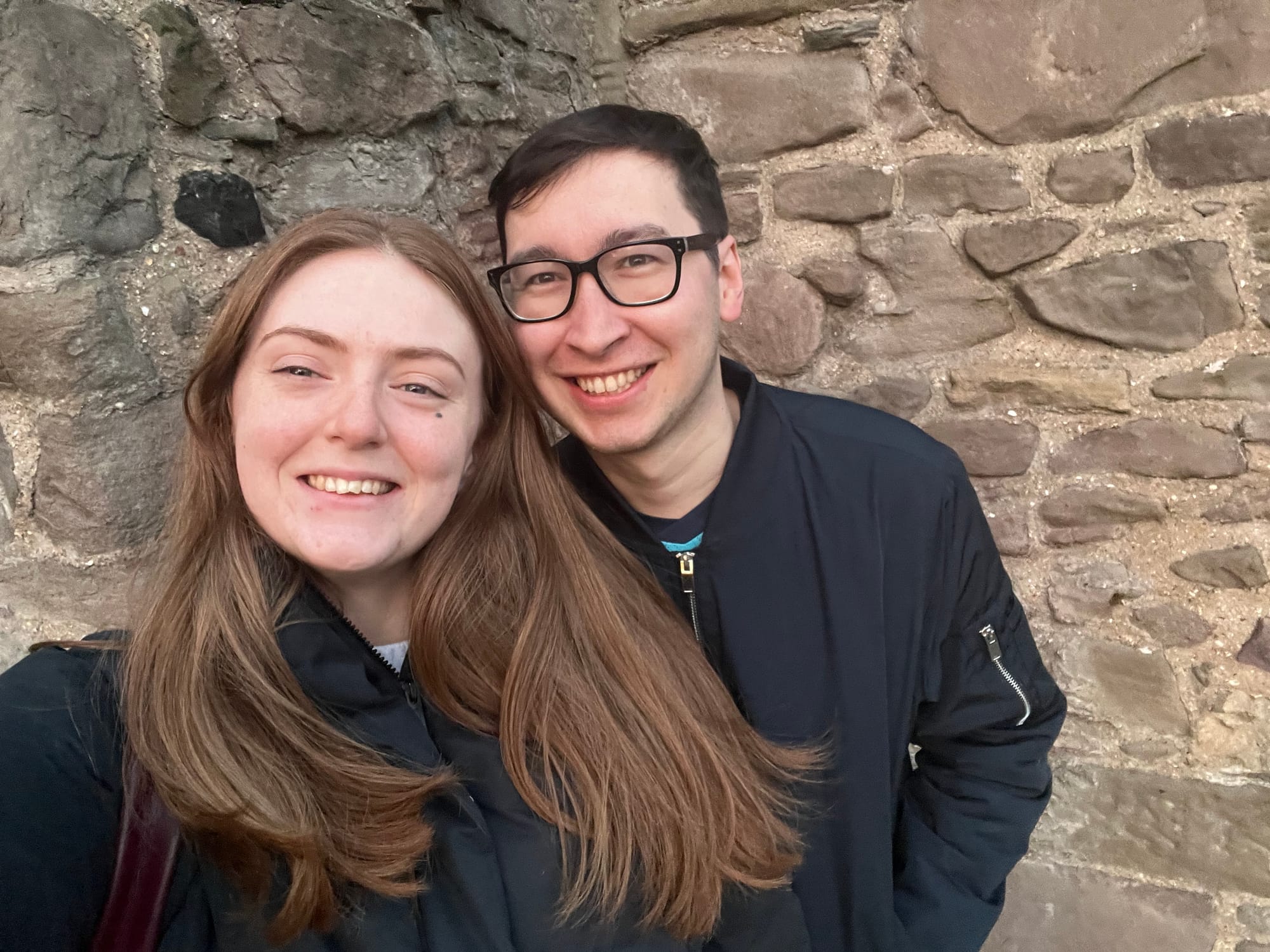 Beth and James smile for a selfie against an old stone wall.