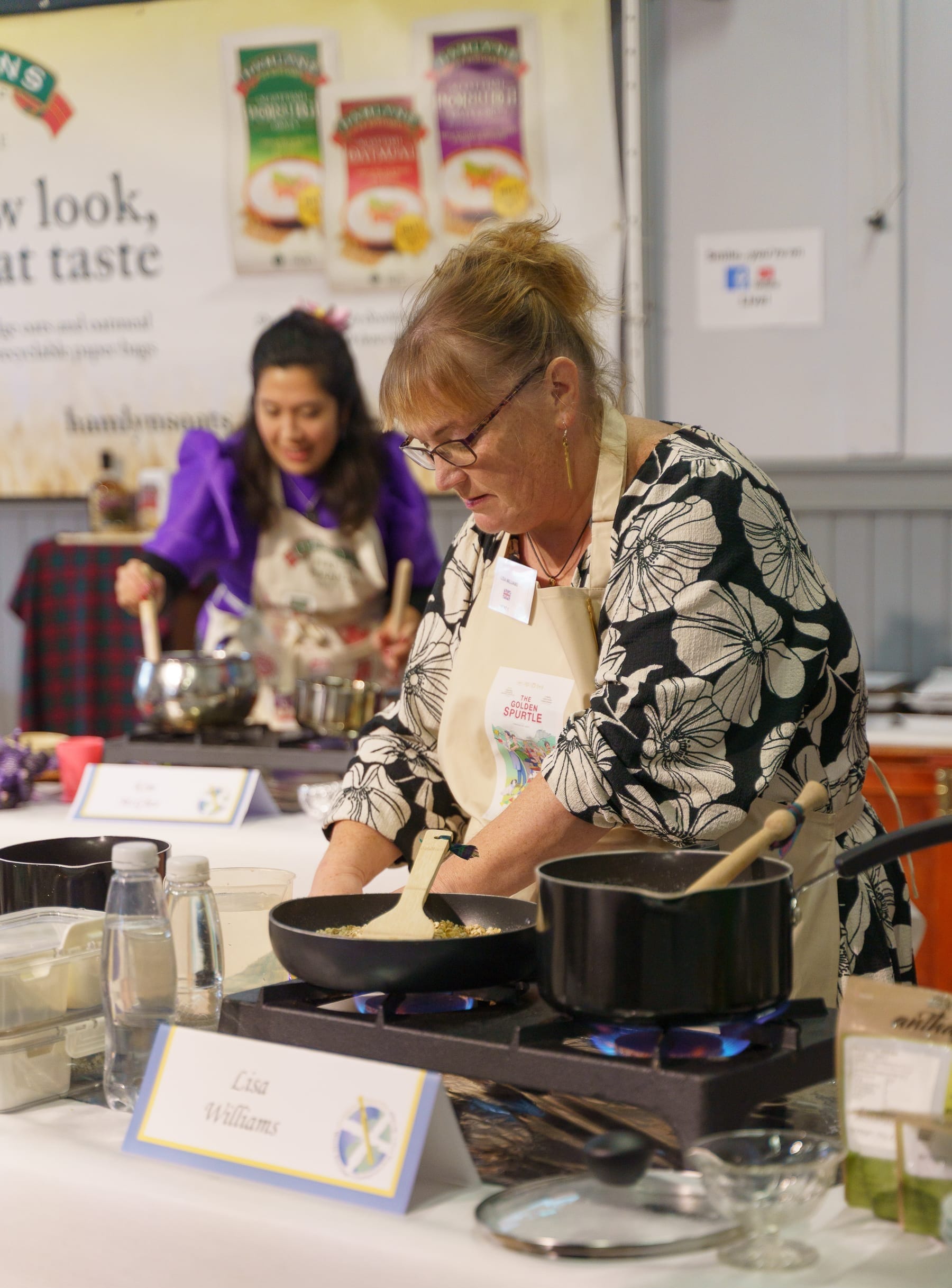 Lisa Williams cooking at her station with pots on the small hob in front of her. She is wearing an apron and concentrating.