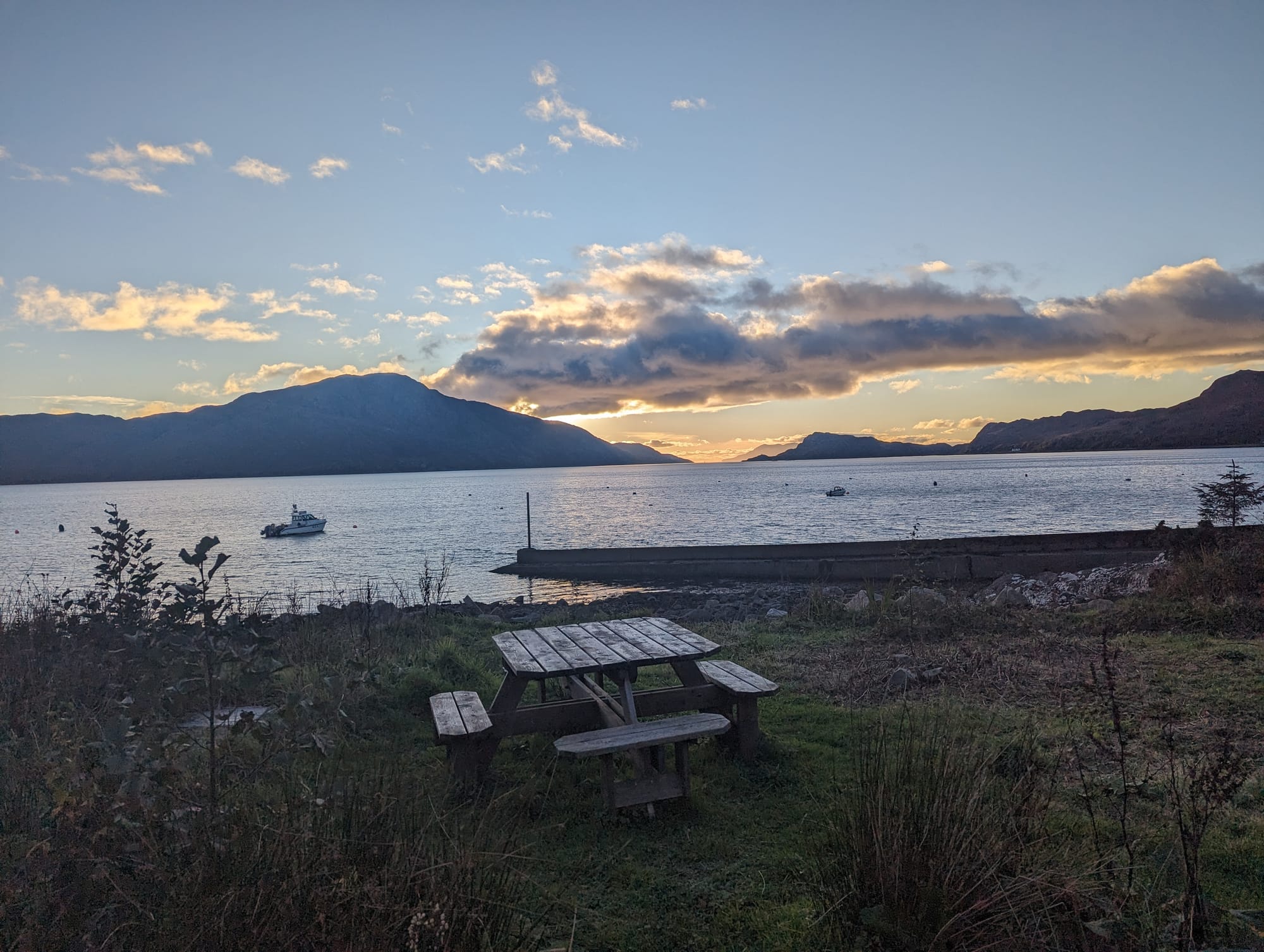 Sunset over Loch Nevis. There is a picnic bench on grass in the foreground.