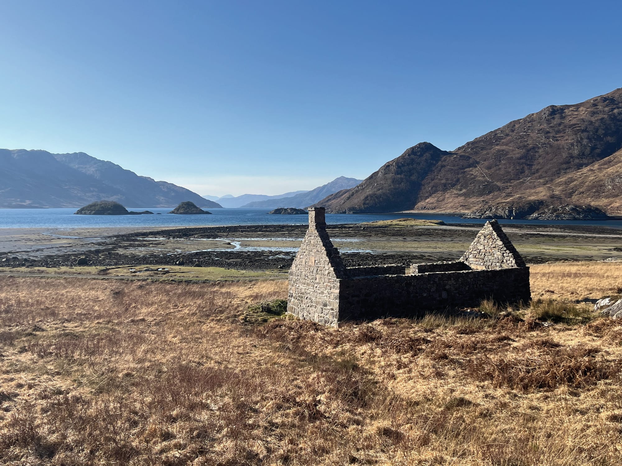 A ruined cottage sits in the foreground with Barrisdale Bay beyond. Blue water and mountains are in the distance.