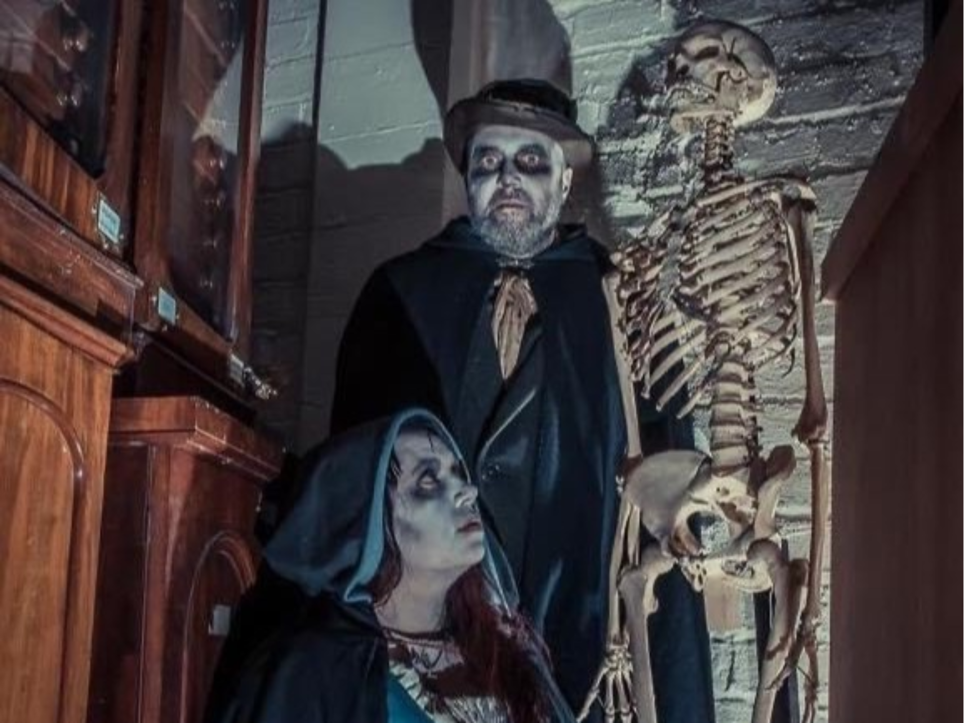 A man and a woman in historic costume next to a human skeleton model. They have their faces painted white and dark make-up around their eyes.