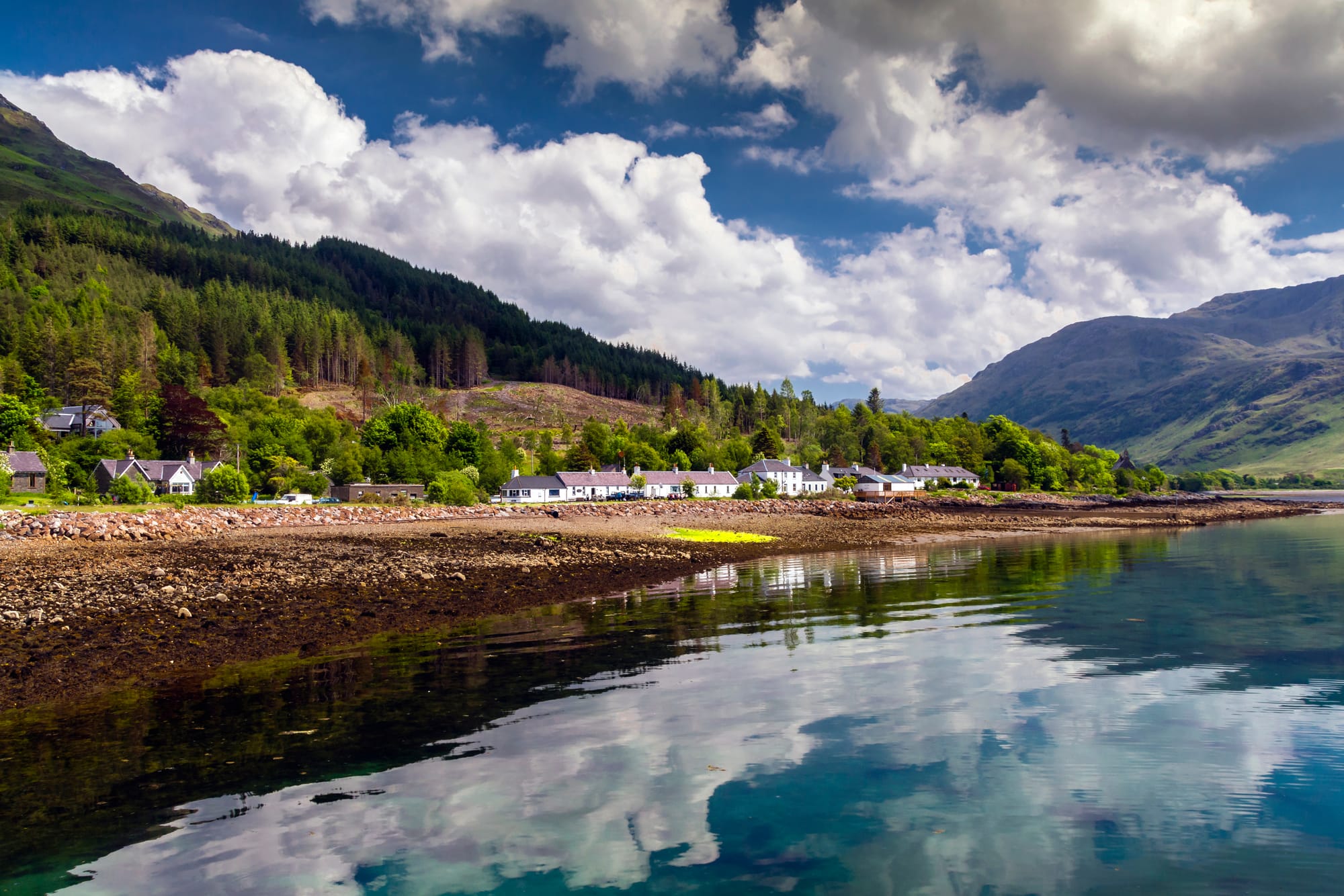 The houses of the village of Inverie sit on the rocky shore along the water. Clouds are reflected in the loch.