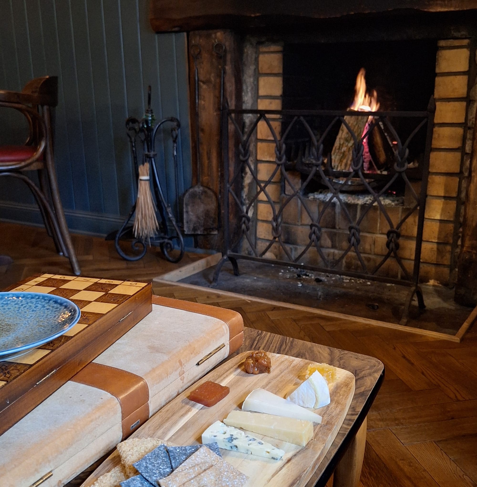 A wooden board with slices of cheese on a coffee table next to a fire.