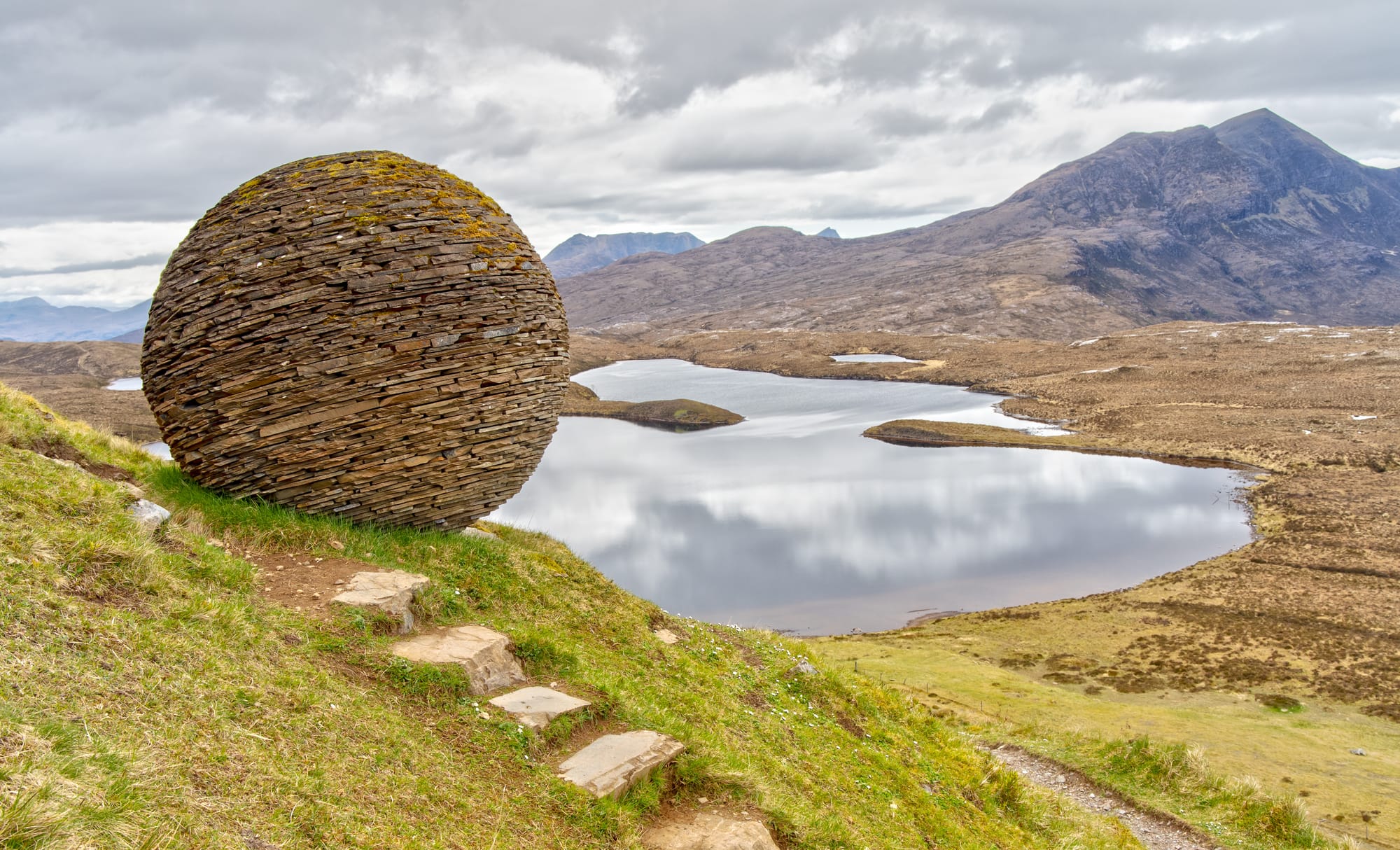 A spherical sculpture made of different textures of rock next to a path alongside a loch.