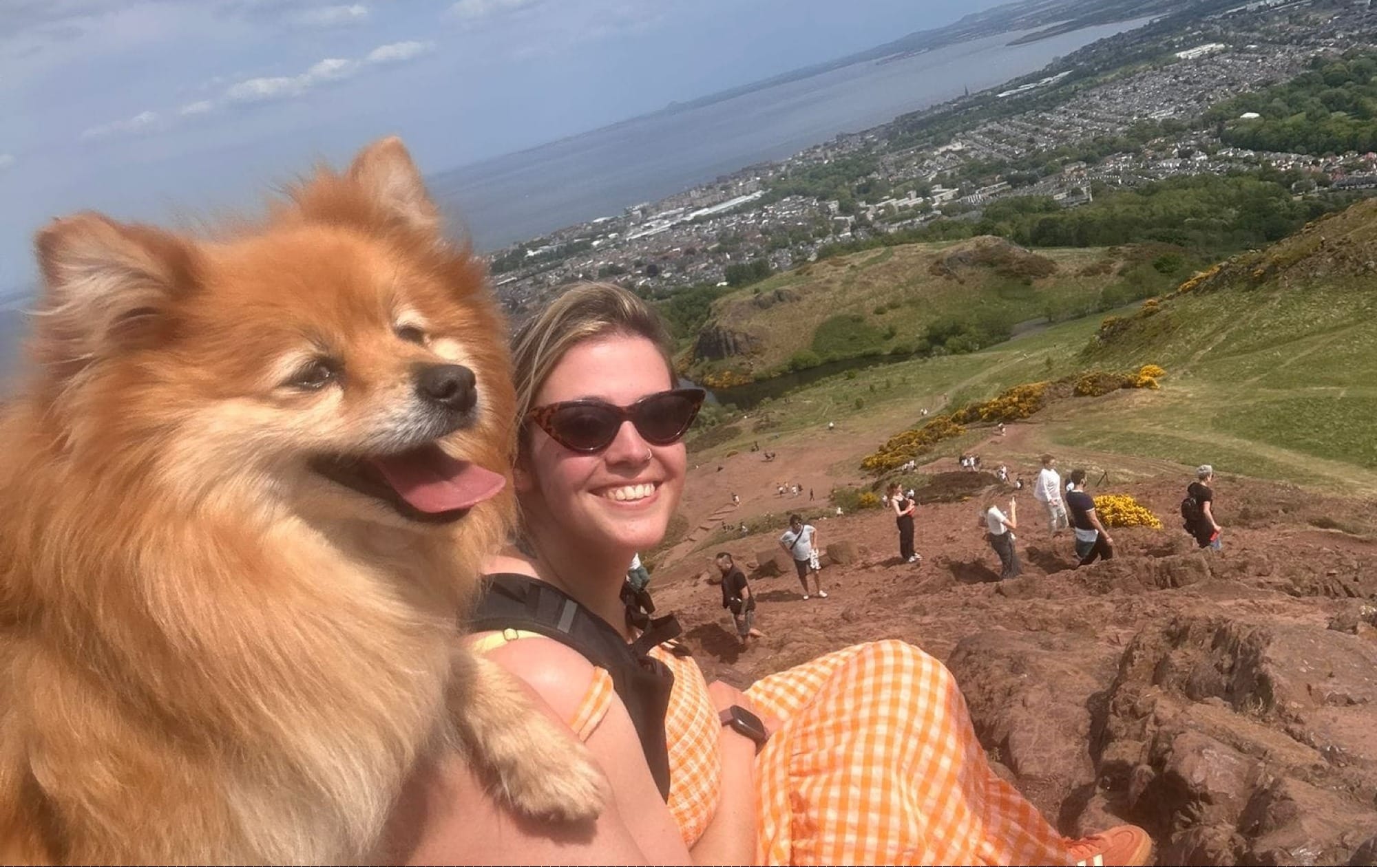 Daniella and Hector the Pomeranian sitting on Arthur's Seat with the city and the sea in the background.
