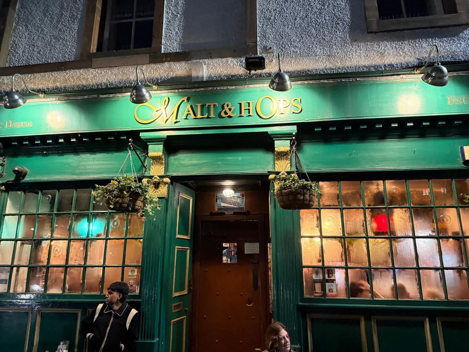 The green-painted exterior of the Malt & Hops. Lights show people inside the pub.