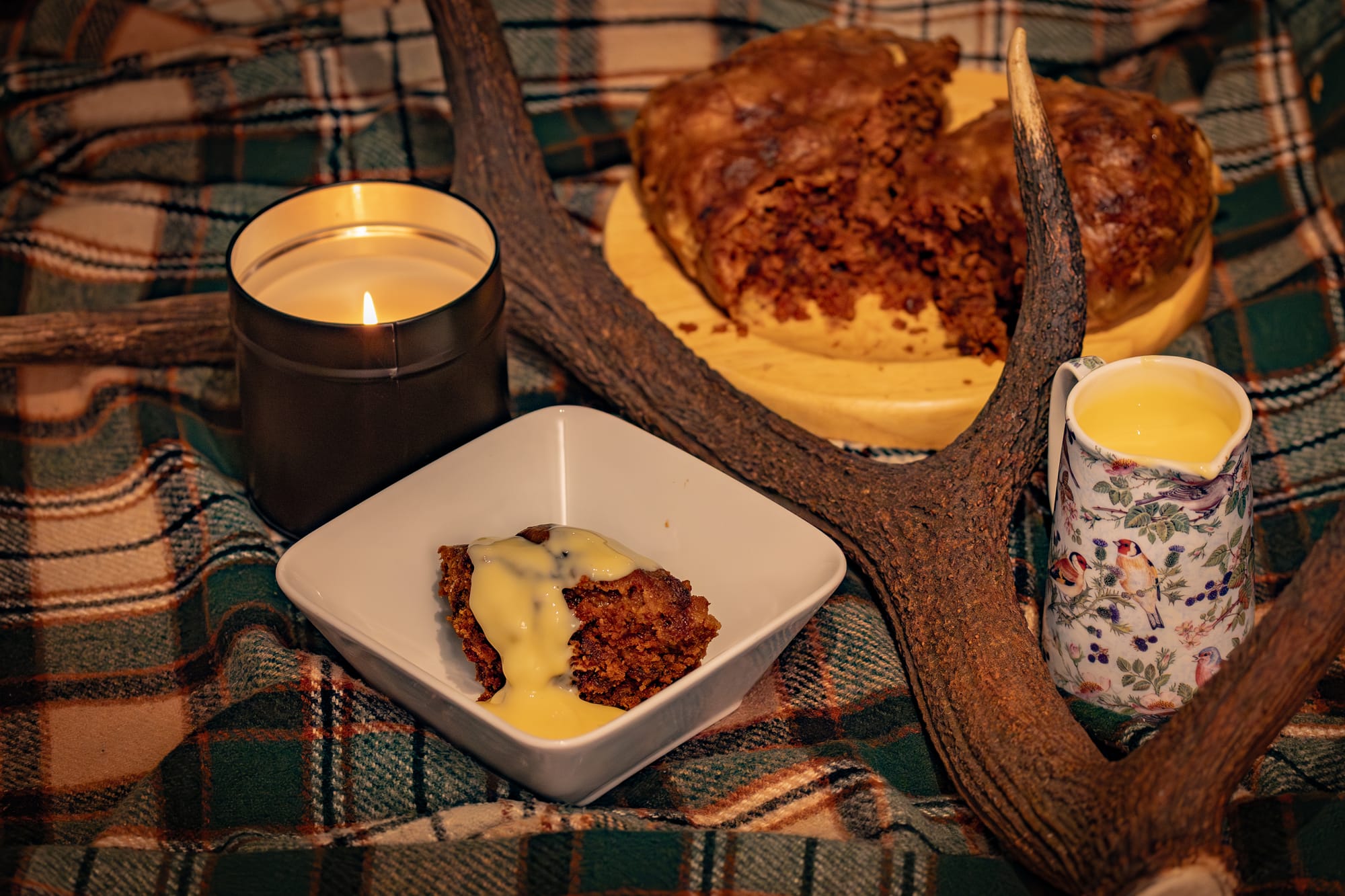 A bowl of clootie dumpling on a tartan blanket. An antler is nearby for decoration and there is a lit candle.
