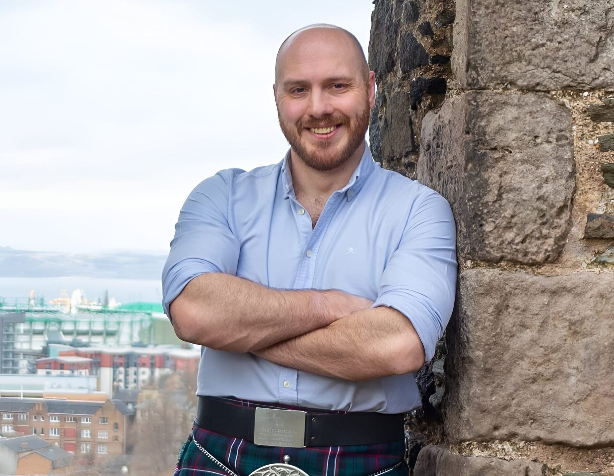 A man in a pale blue shirt with sleeves rolled up and open collar stands smiling at the camera by an old stone wall wearing a greena nd purple kilt.