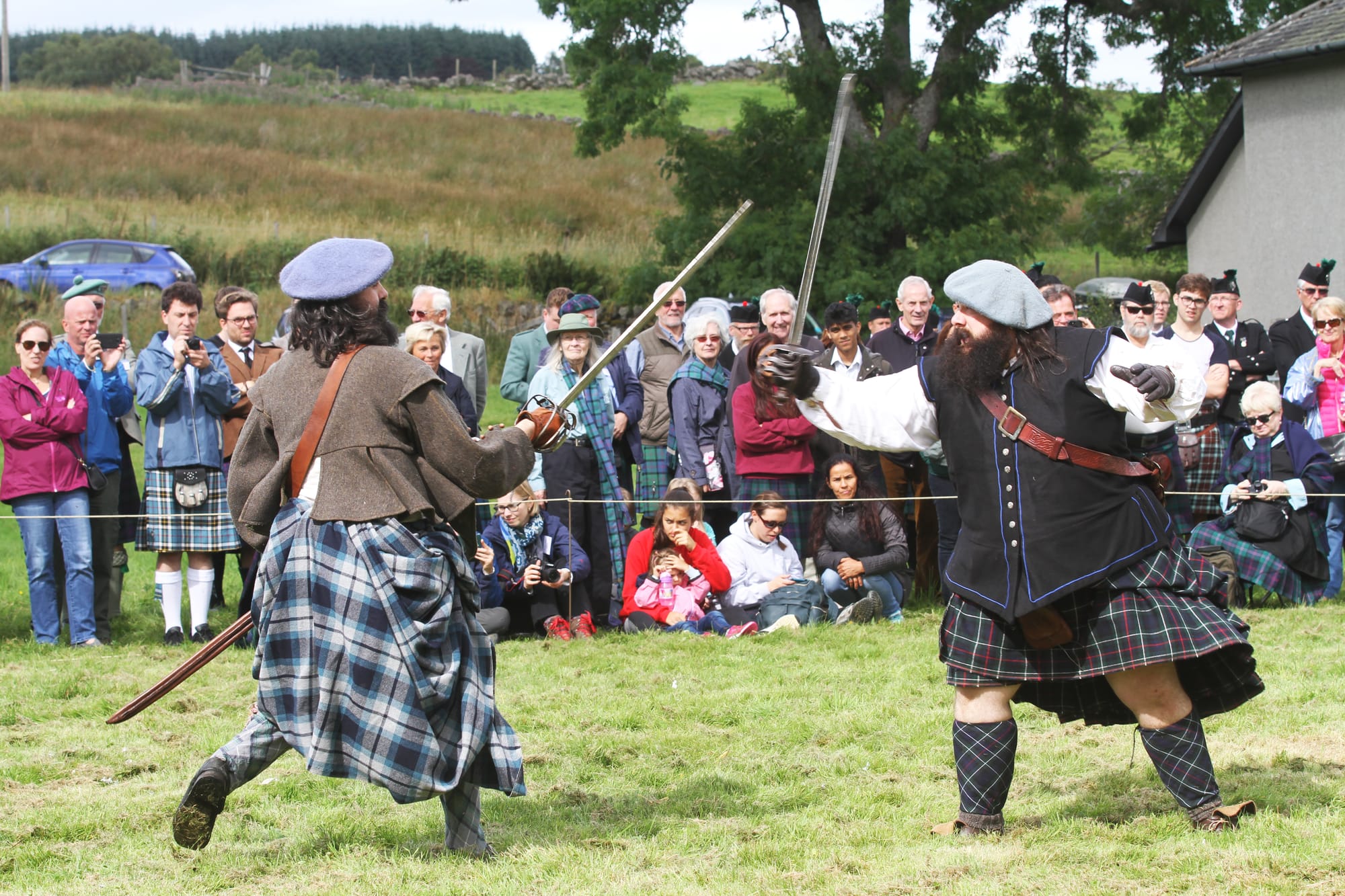 Two men dressed as clansmen re-enact and sword fight while people sit or stand on the grass and watch.