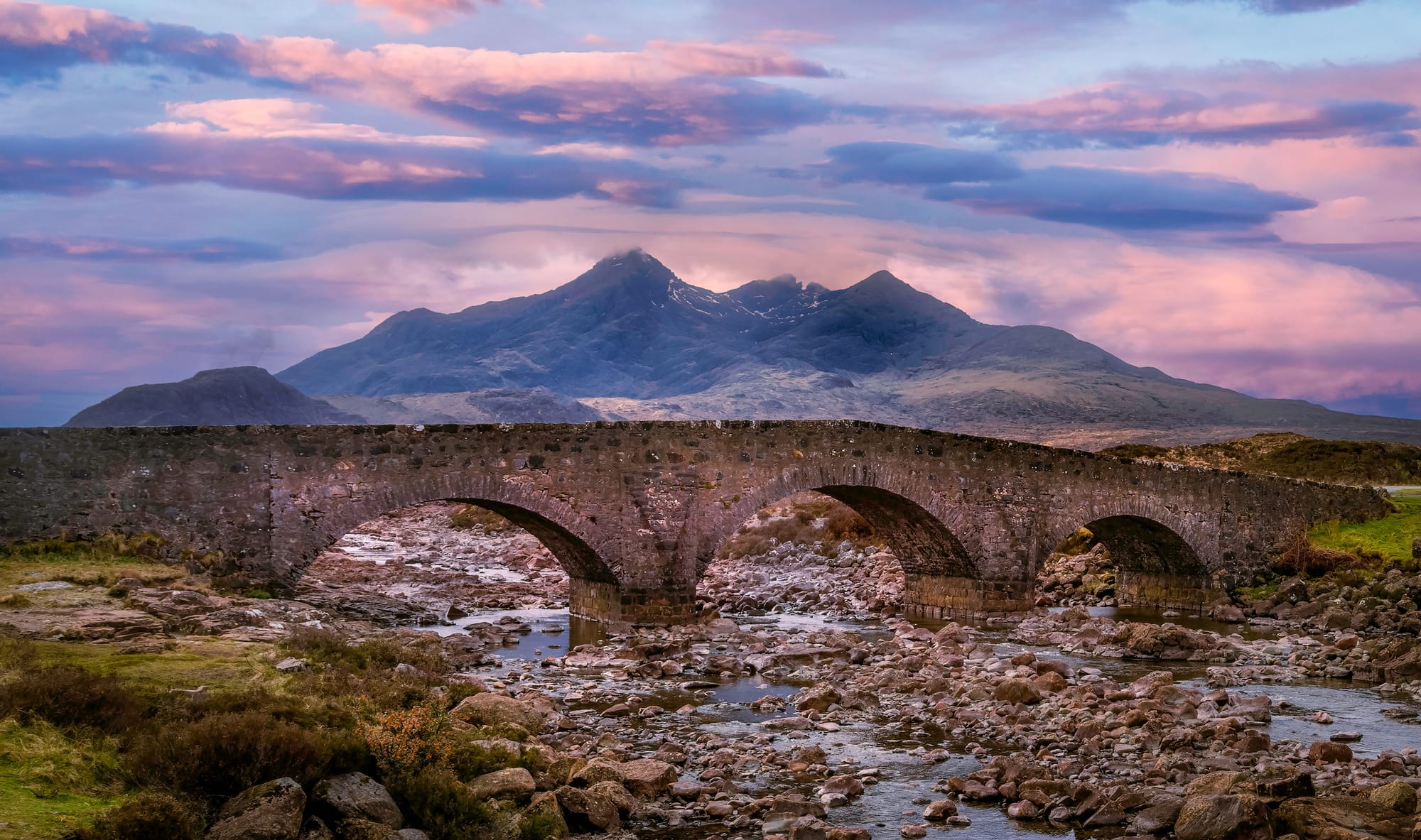 An old stone bridge with a shallow river flowing over large stone below and the spiky Cuillin mountains behind.