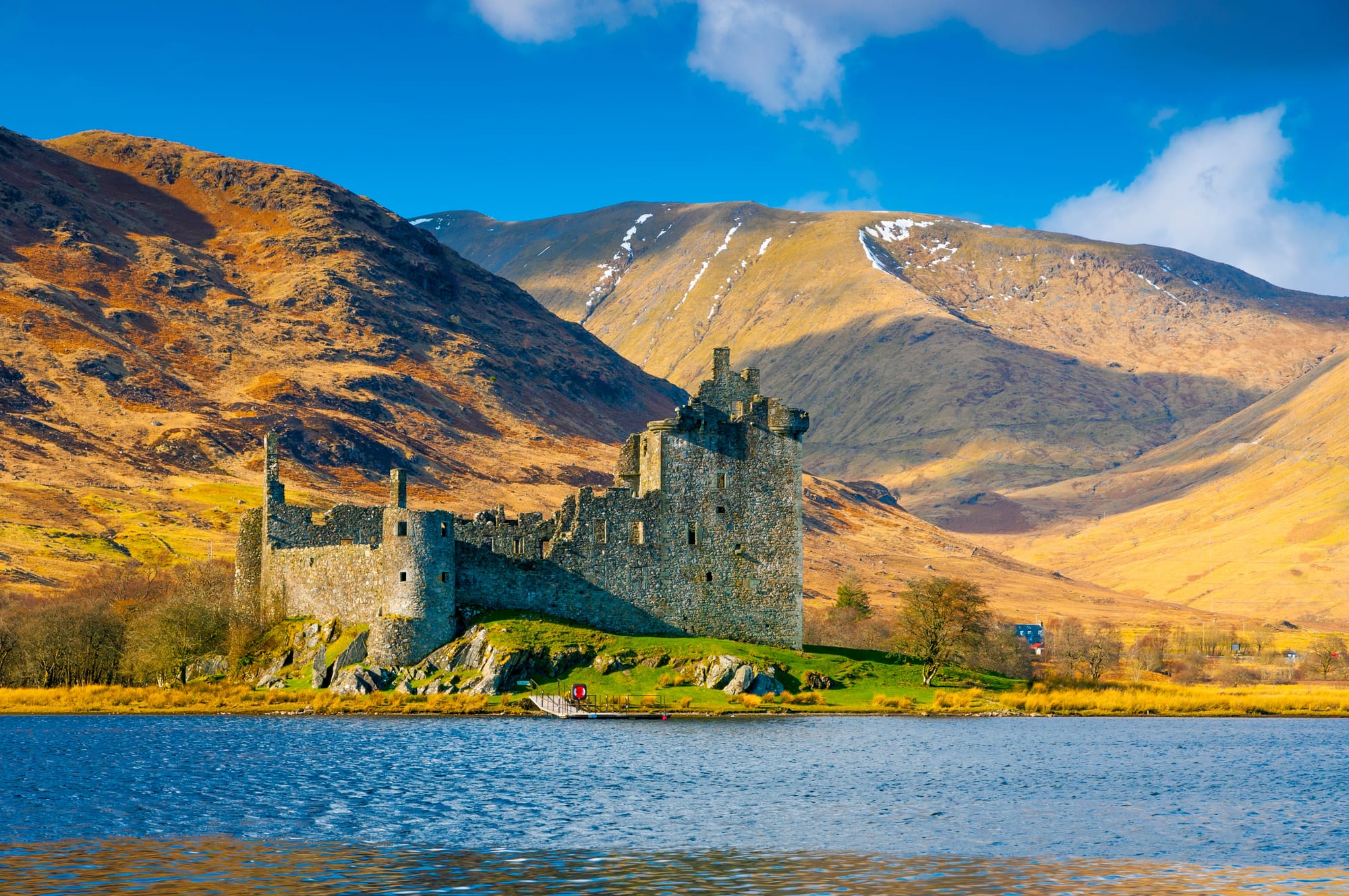A stunning old Scottish castle in ruins on a small peninsula of a loch with large hills behind.