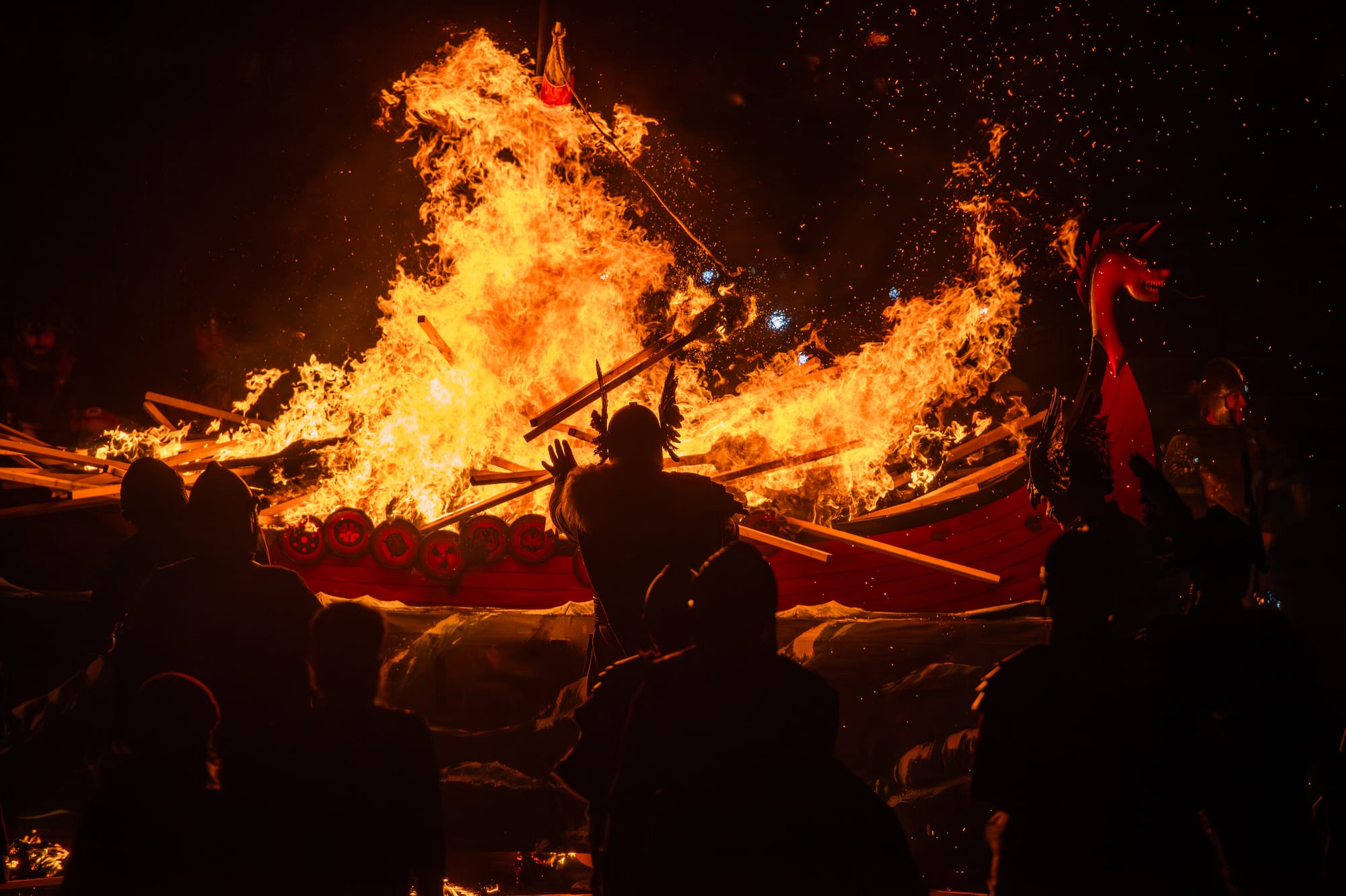 The silhouette of a man in Viking costume standing in front of the galley as it burns.