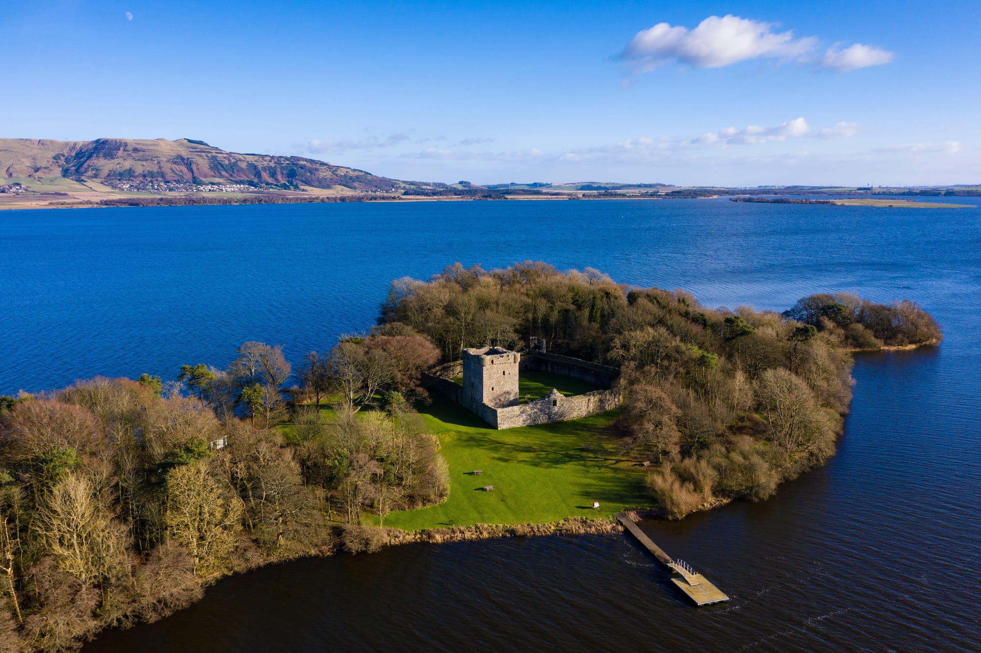 A castle on a small island with a boat jetty in the middle of a blue loch.