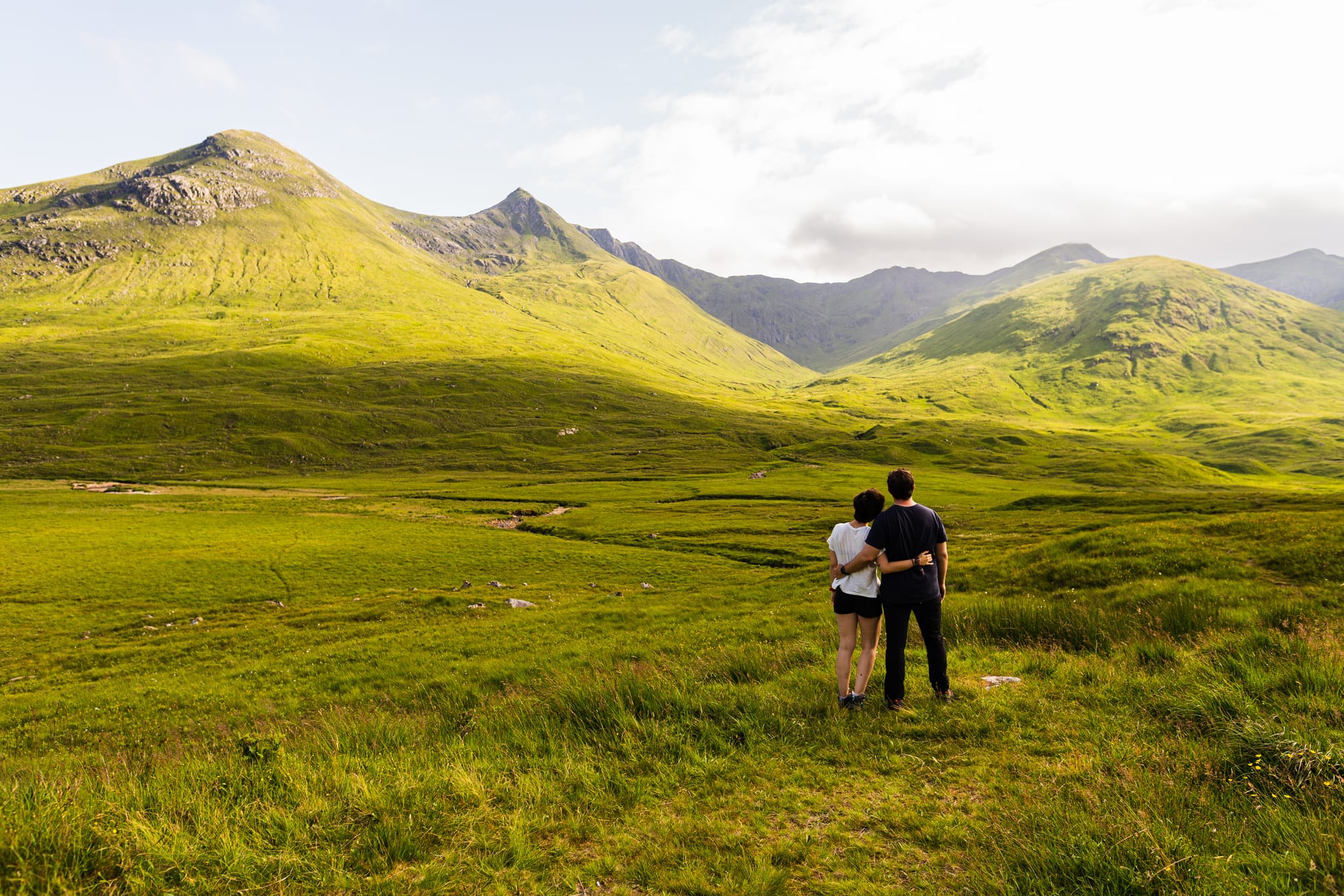 A couple stand with their arms around each other with grassy hills in the background.