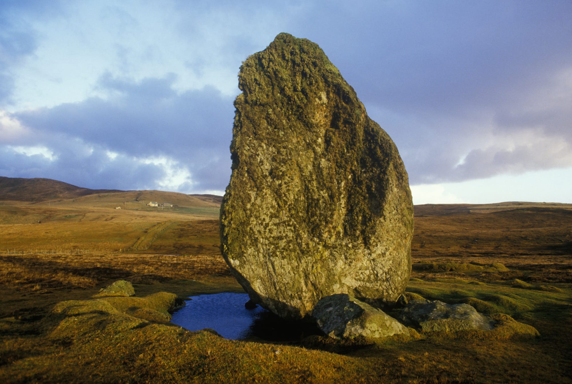 A standing stone in a mossy hill next to a puddle. There are hills in the background. 