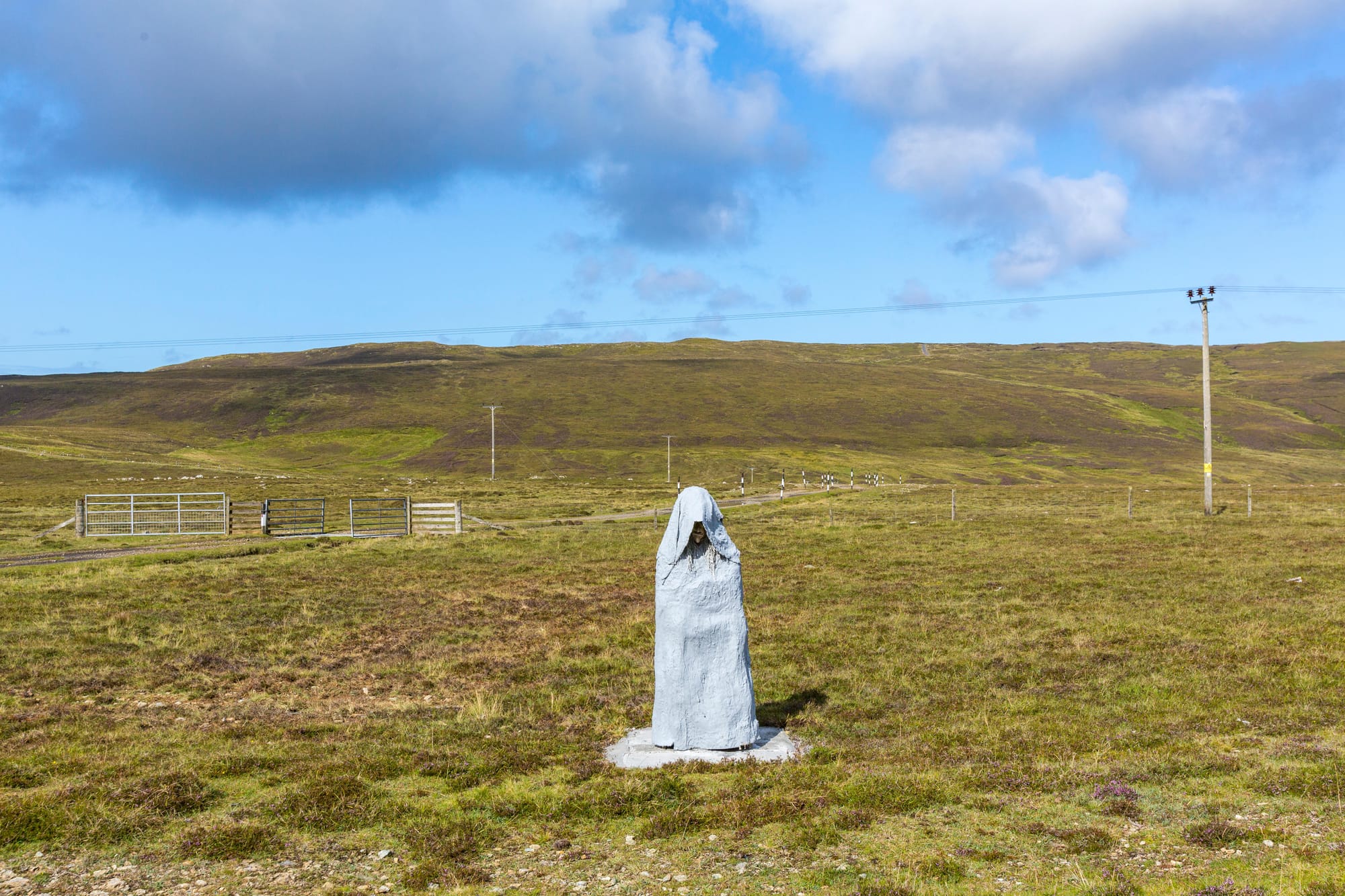 A person-sized statue, designed to look like they are wearing a white cloak wrapped tightly around them. There are green hills in the background.