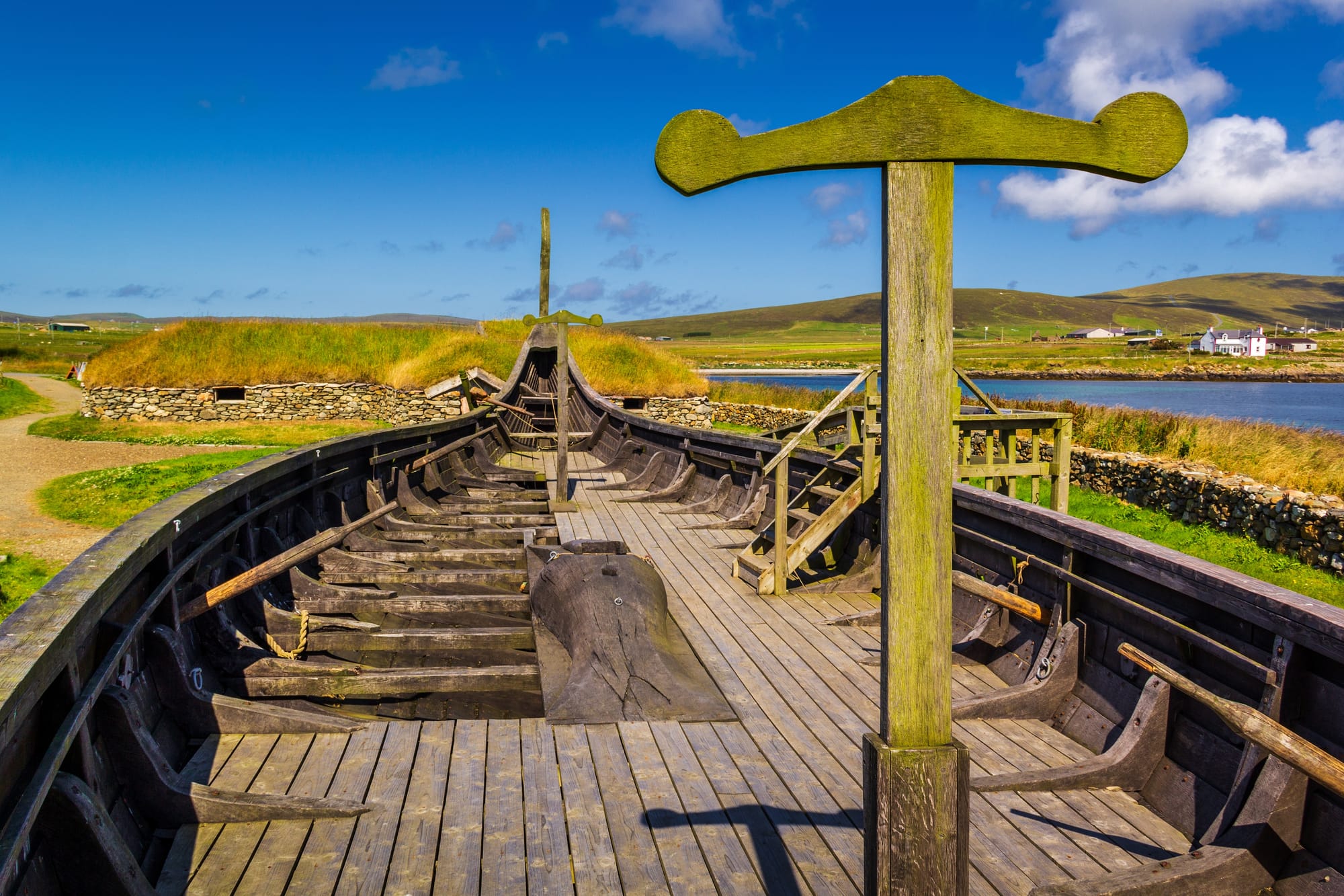 A replica Viking longship made of wood sits on the grassy shore, with water and hills in the background.