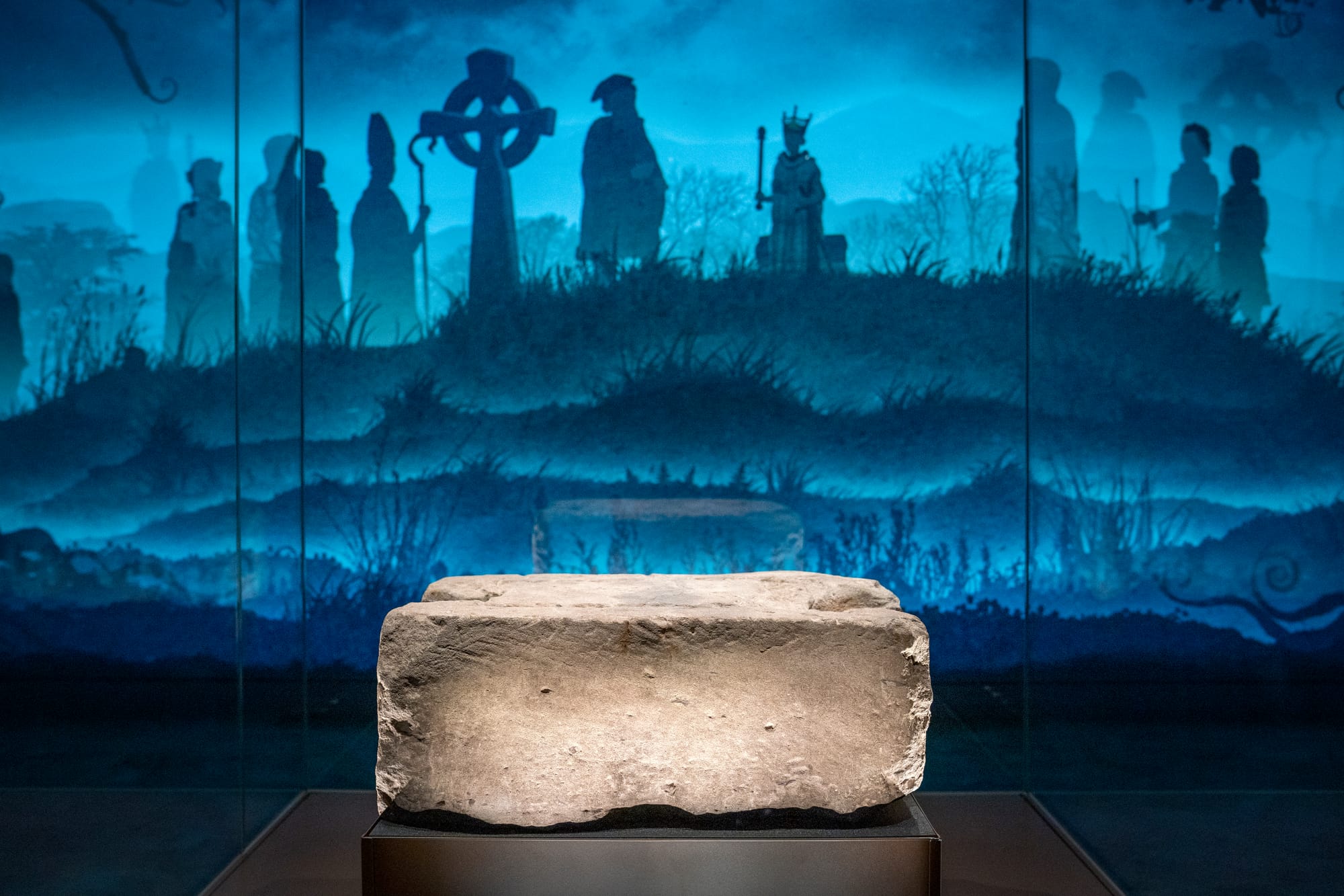 A large grey rectangular stone on a dark coloured raised platform with a bluey background with shadowy figures from Scotland's past.