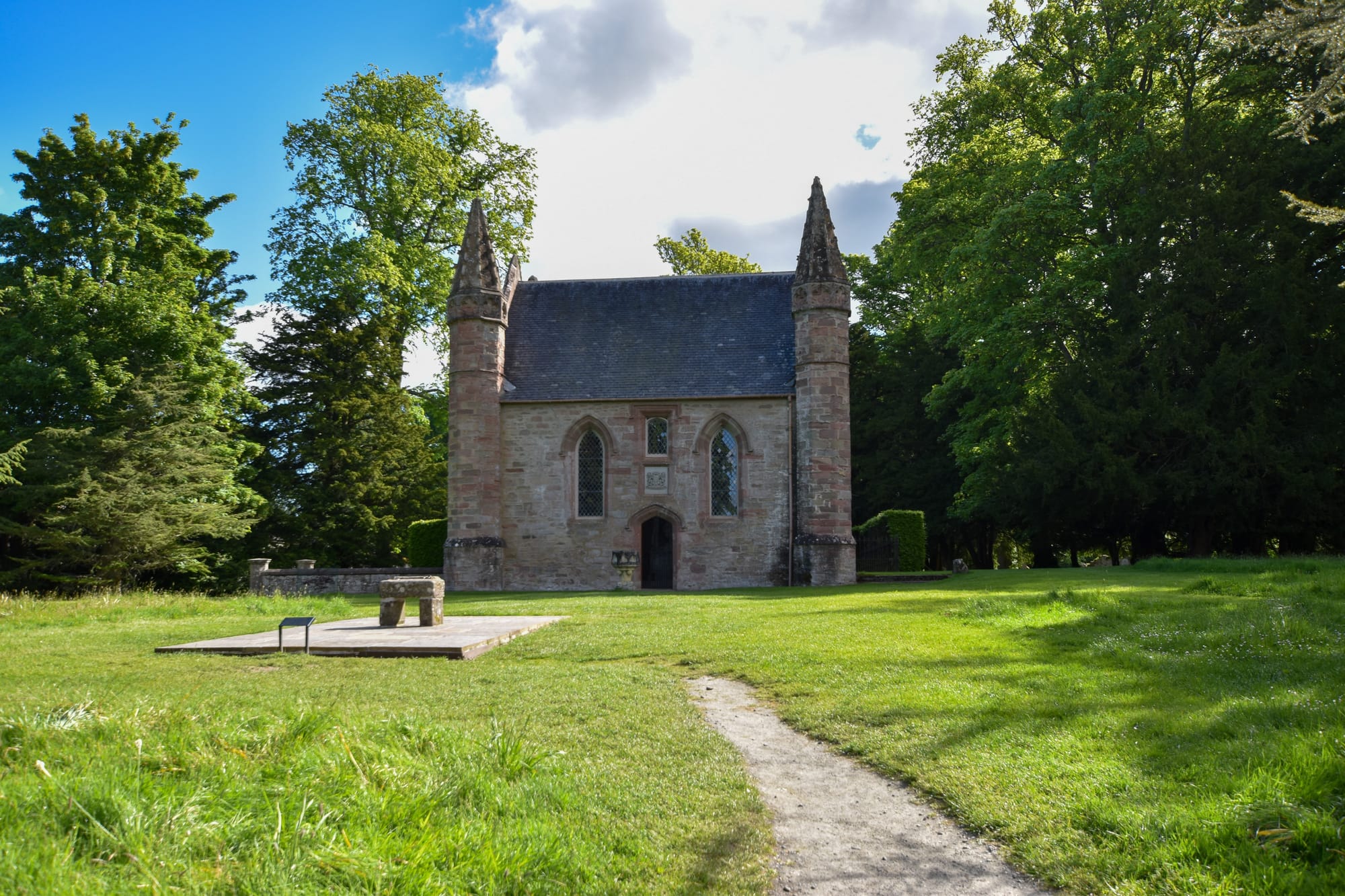 A small simple stone chapel with pointed turrets at each four corners and a replica of the stone of destine out front on a stone slab among well-maintained lawn.