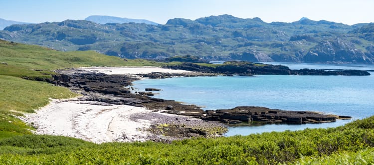 A beautiful white sand beach with blue water and surrounded by rugged hills and fields.