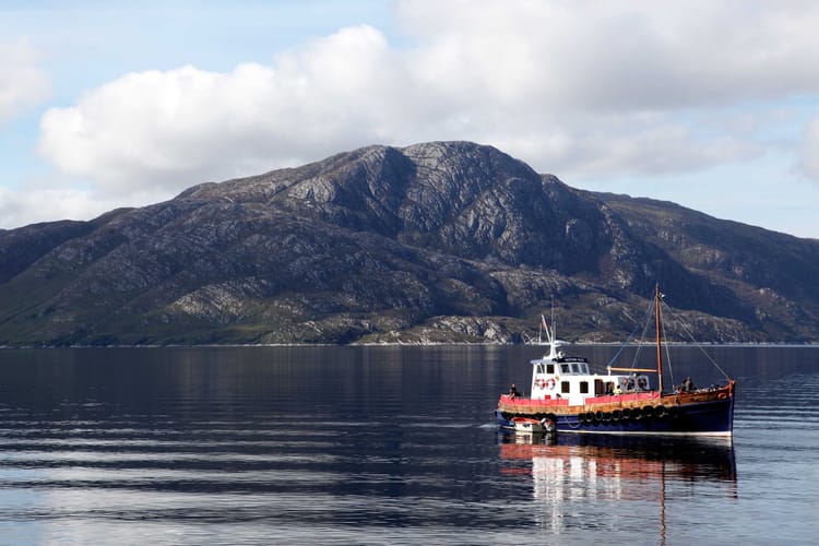 The small white, red and blue ferry sailing from Mallaig to Knoydart. There is a rocky mountain in the background