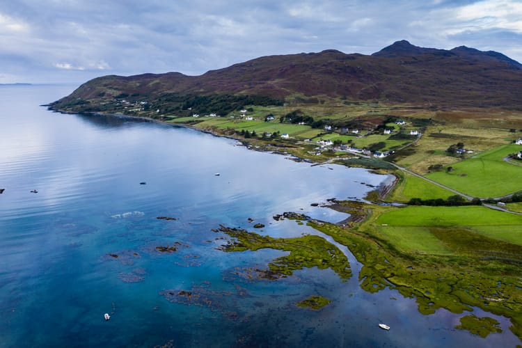 Houses dotted along the coast of the Ardnamurchan peninsula. The water is very blue and the land is green, with heather-covered mountains in the background.