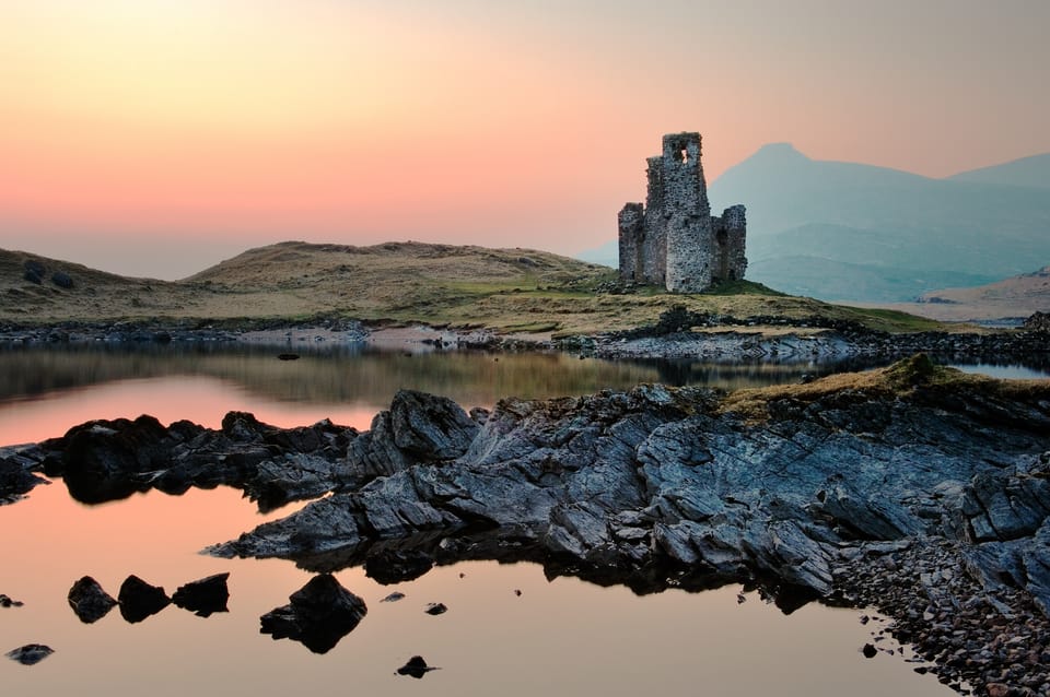 The ruined Ardvreck Castle. In the foreground there are rocks and a loch, and the sky is pink and yellow.