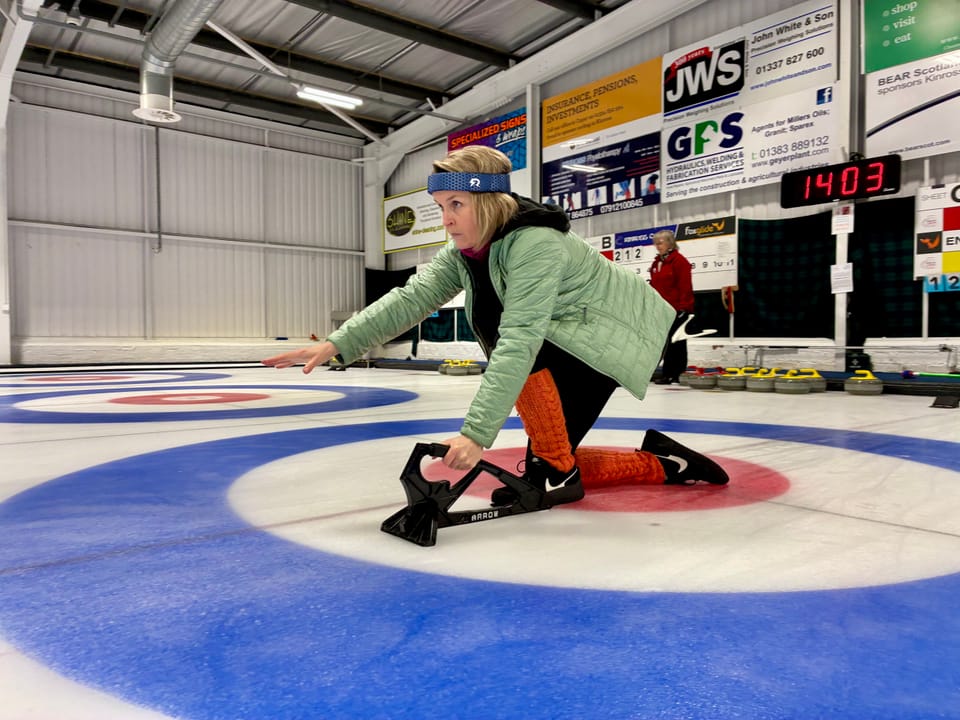 A woman with short blond hair wearing a blue headband and a green jacket is crouched on the ice of a curling rink with arm outstretched after releasing a curling stone.