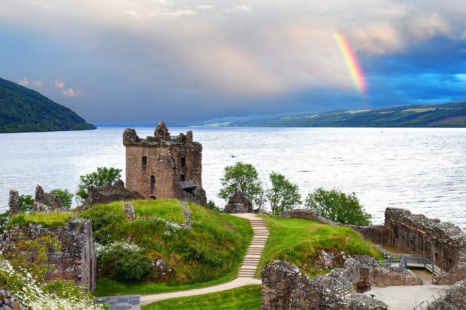 Urquhart Castle on the banks of Loch Ness. There is a rainbow in the background and the water is silvery. 
